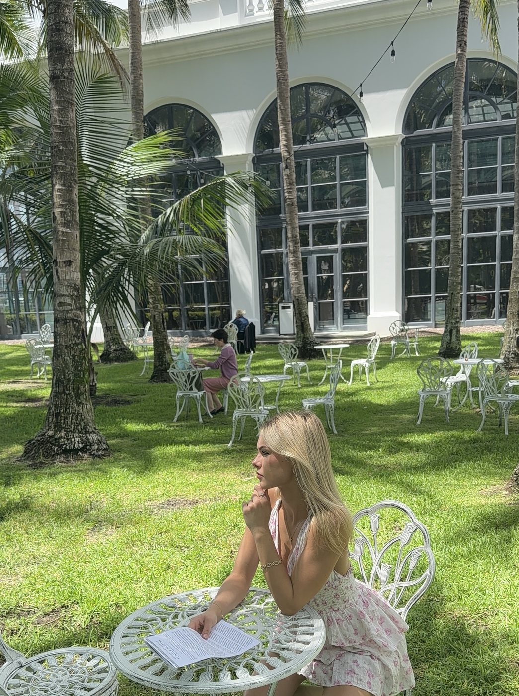 Madeline reading at a white garden table under palm trees